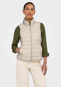 Gilet léger beige en duvet avec un col haut, fermeture éclair sur le devant et poches latérales, porté sur une chemise vert foncé et un pantalon clair.