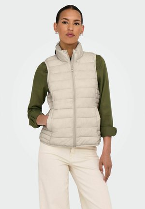 Chaleco acolchado de color beige claro con cuello alto, cierre con cremallera frontal y bolsillos laterales, llevado sobre una camisa verde oscura y pantalones claros.