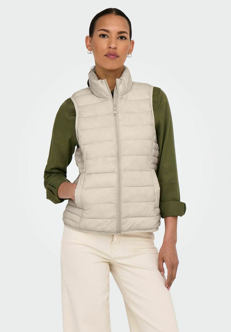 Gilet léger beige en duvet avec un col haut, fermeture éclair sur le devant et poches latérales, porté sur une chemise vert foncé et un pantalon clair.