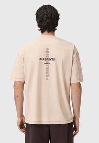Camiseta de manga corta beige con un corte holgado. La parte de atrás presenta letras verticales en negro que dicen "ALLSAINTS LONDON" con un acento texturizado en rosa.