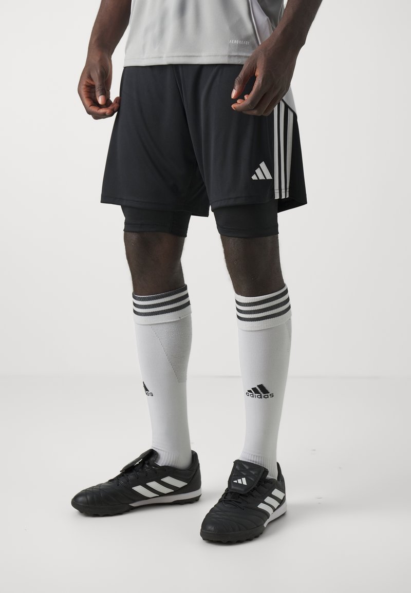Athlète portant un short noir Adidas, des chaussettes hautes blanches Adidas avec des rayures noires et des chaussures de football Adidas noires, debout sur un sol blanc.