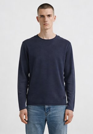 Giovane uomo con capelli corti che indossa una camicia blu navy a maniche lunghe e jeans blu, in piedi davanti a uno sfondo chiaro e semplice.