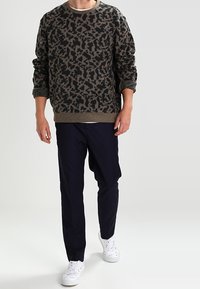 Maglione a righe marroni e nere con maniche lunghe, abbinato a pantaloni blu e sneakers bianche. Il materiale appare morbido con una finitura testurizzata.