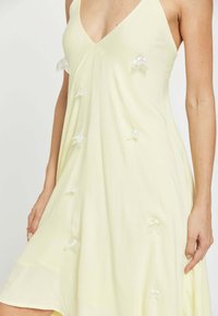 Mujer con vestido amarillo pálido sin mangas con adornos transparentes en forma de pétalo esparcidos por el frente.
