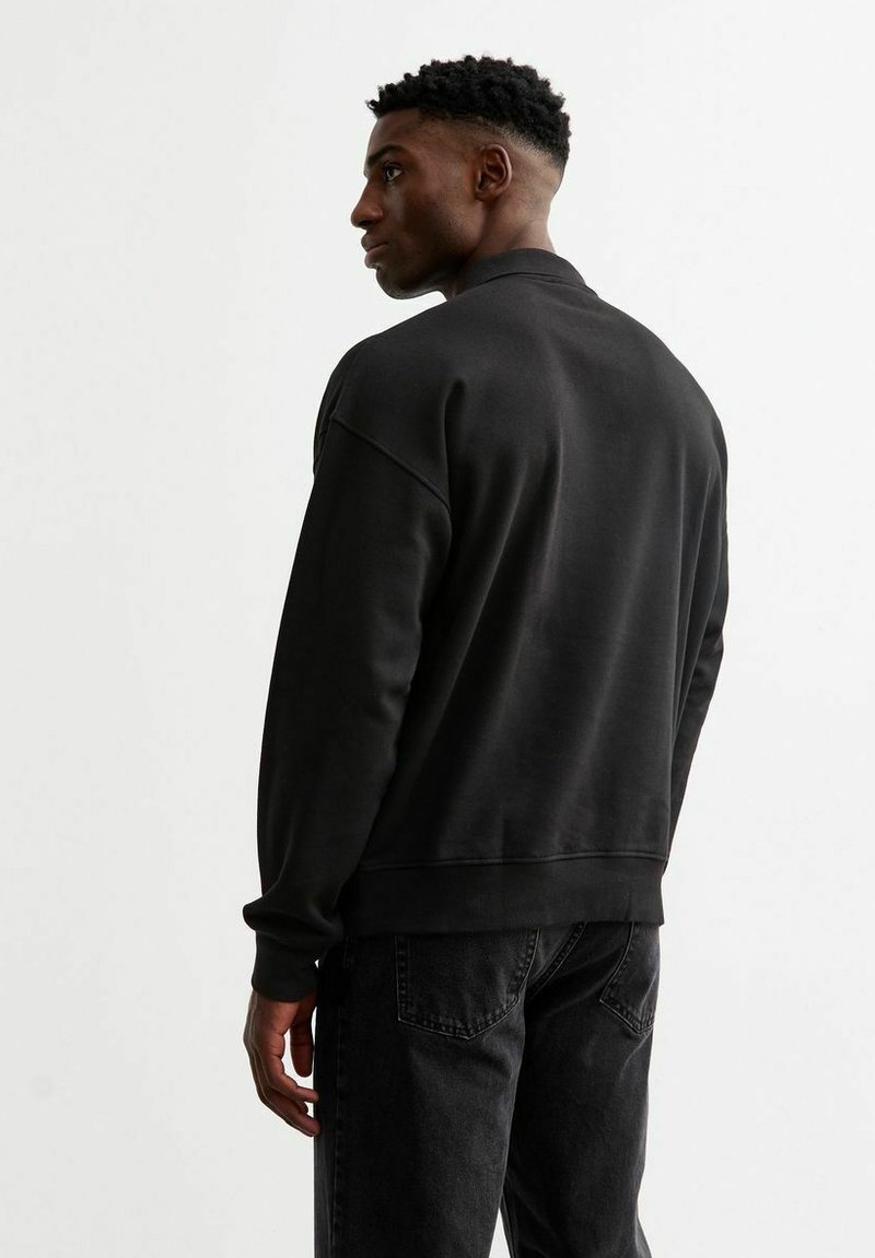 Felpa nera con vestibilità rilassata e polsini a coste. Tessuto liscio, design semplice e scollatura arrotondata. Indossata con jeans scuri.
