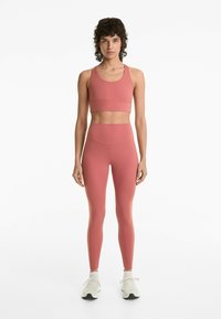 Rosa sport-bh och högmidjade leggings i slätt, stretchy tyg. Modellen bär vita sneakers. Enkelt, figurnära design.