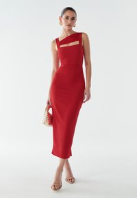 Rød tætsiddende midi-kjole med asymmetrisk stropdesign og nøglehuldsdetalje. Glat stof med en bodycon pasform, kombineret med en vævet håndtaske.