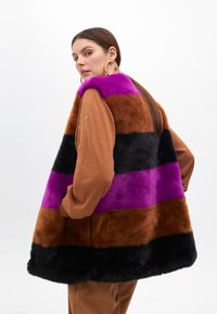 Colorful faux fur vest featuring horizontal stripes in purple, brown, black; paired with a brown long-sleeve top and gold hoop earrings.