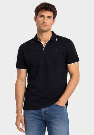 Mann trägt schwarzes kurzärmeliges Poloshirt mit weißem Besatz und blaue Jeans, steht mit einer Hand in der Tasche vor schlichtem Hintergrund.