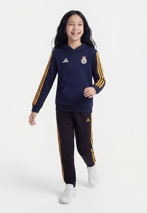 Niña sonriente caminando, vestida con sudadera con capucha azul marino y pantalones negros con rayas naranjas, zapatillas blancas, con los logotipos del Real Madrid y Adidas.
