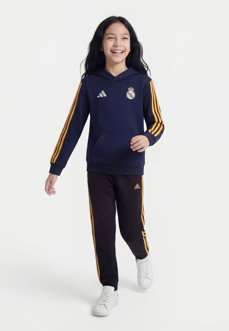 Fille souriante marchant, portant un sweat à capuche bleu marine et un pantalon noir avec des bandes orange, des baskets blanches, ainsi que les logos du Real Madrid et d'Adidas.