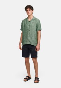 Camicia verde in lino a maniche corte con colletto, bottoni frontali e un piccolo logo; abbinata a pantaloni corti neri e sandali scuri.