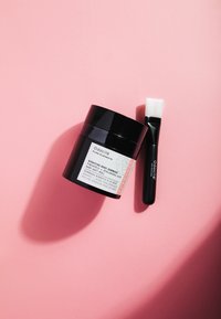 Pot noir de produit de soin de la peau avec une étiquette blanche, accompagné d'un pinceau noir avec une pointe de poils blancs, reposant sur un fond rose.