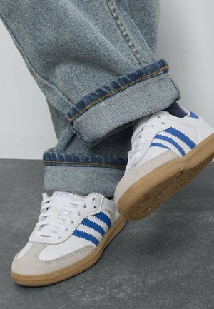 Chaussures de sport blanches avec des rayures bleues, des détails en daim et une semelle en caoutchouc. Associées à un jean en denim clair retroussé.