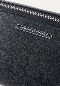Crni teksturirani kožni novčanik s srebrnim metalnim zatvaračem i utisnutom pločicom s logotipom "Armani Exchange" sprijeda.