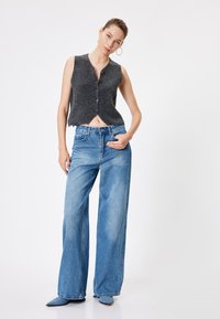 Charcoalgrijs gebreid vest met een diepe V-hals en knopen aan de voorkant, gecombineerd met lichtblauwe wijde jeans. De stof oogt zacht met een zichtbare textuur.