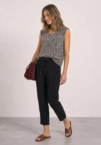 Woman standing in black drawstring pants, leopard print sleeveless top, brown sandals, and holding a dark red handbag against a plain wall.