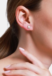 Boucles d'oreilles en argent en forme de fleur avec des accents de gemmes blanches, montées sur un fond doré, portées sur l'oreille d'un modèle. Peau lisse.