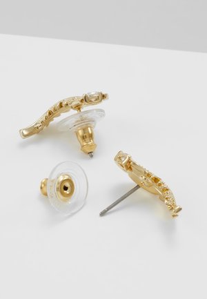 Paire de boucles d’oreilles à clous en métal doré avec attaches en plastique transparent, ornées de petits cristaux incrustés sur une surface blanche.