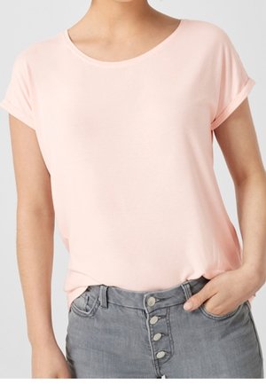 Femme portant une chemise rose clair à manches courtes rentrée dans un jean gris taille haute avec quatre boutons visibles, main dans la poche.