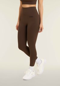 Leggings a vita alta marroni con una texture elastica e liscia, dotati di un piccolo logo nero. Abbinati a sneakers bianche e calzini corti bianchi.