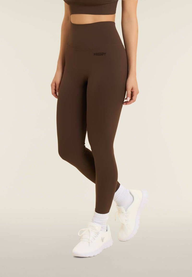 Leggings a vita alta marroni con una texture elastica e liscia, dotati di un piccolo logo nero. Abbinati a sneakers bianche e calzini corti bianchi.
