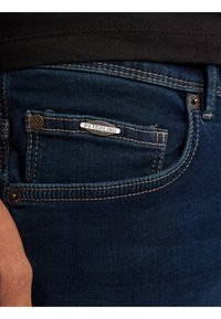 Jeans denim w ciemnoniebieskim kolorze, z małą przednią kieszenią z srebrną etykietą "PETROLIND" i kontrastującymi detalami ściegów.