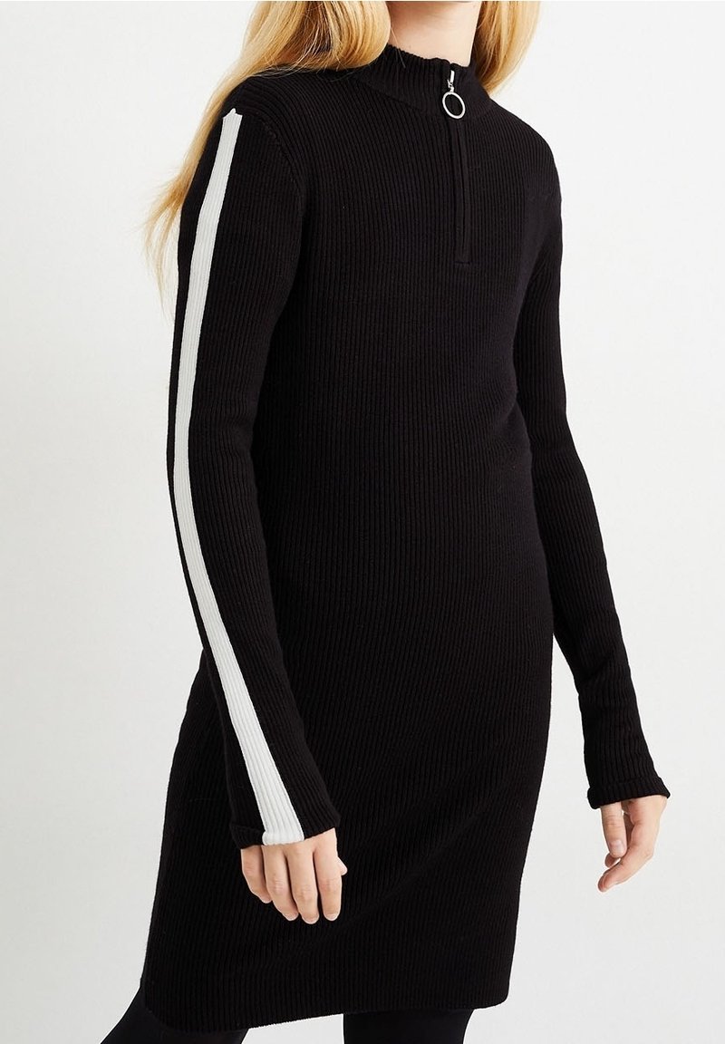 Femme portant une robe noire côtelée à manches longues, avec une bande blanche sur la manche et une fermeture éclair frontale avec un tire-zip circulaire.