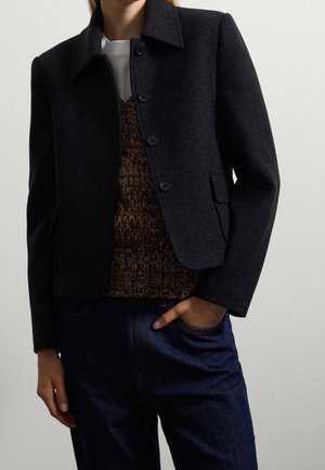 Person wearing buttoned black wool jacket, brown knitted vest, white shirt, and dark blue jeans with one hand in pocket.