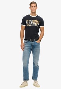 Camiseta negra de manga corta con ribete blanco que presenta un diseño de logo en capas. Combinada con jeans de mezclilla azul y zapatillas beige. Fondo liso.