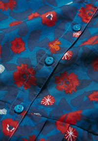 Niebieska tkanina w kwiatowy wzór z czerwonymi i białymi kwiatami. Posiada małe niebieskie guziki oraz teksturowany wygląd z zakładkami wzdłuż krawędzi.