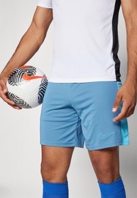 Nike futbolo šortai šviesiai mėlyni su turkio akcentais, derinami su balta marškinėliais ir mėlynomis kojinėmis, laikant raštuotą futbolo kamuolį.