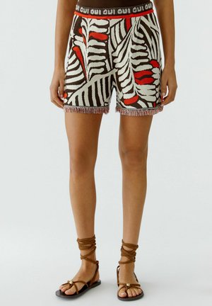 Bruine, witte en rode gebreide shorts met patroon, franjes aan de zoom, elastische tailleband met tekst "oui", gedragen met bruine veter-sandalen op blote benen.