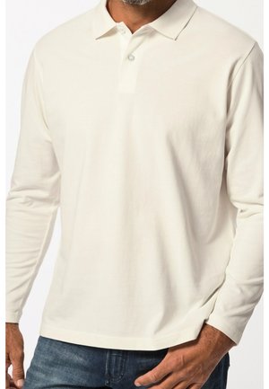 Mand iført en langærmet, off-white polo med knapper, kombineret med blå jeans, hånd hvilende nær lommen.