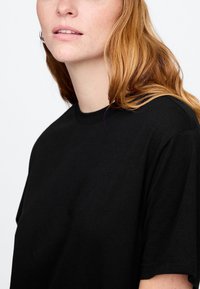 Črna majica s kratkimi rokavi iz bombaža, s klasičnim okroglim ovratnikom in gladko teksturo, prikazuje podrobnosti ramena in vratu.