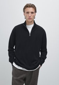 Zwarte zip-up sweater van zachte stof met een hoge kraag, gecombineerd met een wit t-shirt eronder en gestructureerde grijze broek.