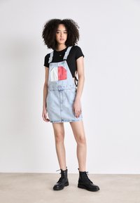 Vestido de tirantes de mezclilla en azul claro con un parche cuadrado blanco y rojo. Combinado con una camiseta negra y botas negras con cordones. Textura suave.
