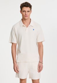Polo shirt beige clair en tissu doux avec des manches courtes, comportant un logo bleu sur la poitrine à gauche, associé à un short assorti.