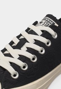 Μαύρο αθλητικό παπούτσι Converse με λευκά κορδόνια, μεταλλικές οπές και μπεζ σόλα, που δείχνει κοντινό πλάνο του δέσιμου και του λογότυπου στη γλώσσα.