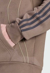 Hnedá mikina s krémovým lemovaním, s tromi tmavými pruhmi na rukáve a klokannou vreckom, vyrobená z textúrovanej látky.
