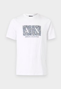 Άσπρο κοντομάνικο T-shirt με μπλε λουλουδάτα μοτίβα σε μπλοκ που περιέχουν τα γράμματα "A" και "X" πάνω από το κείμενο "Armani Exchange" μπροστά.