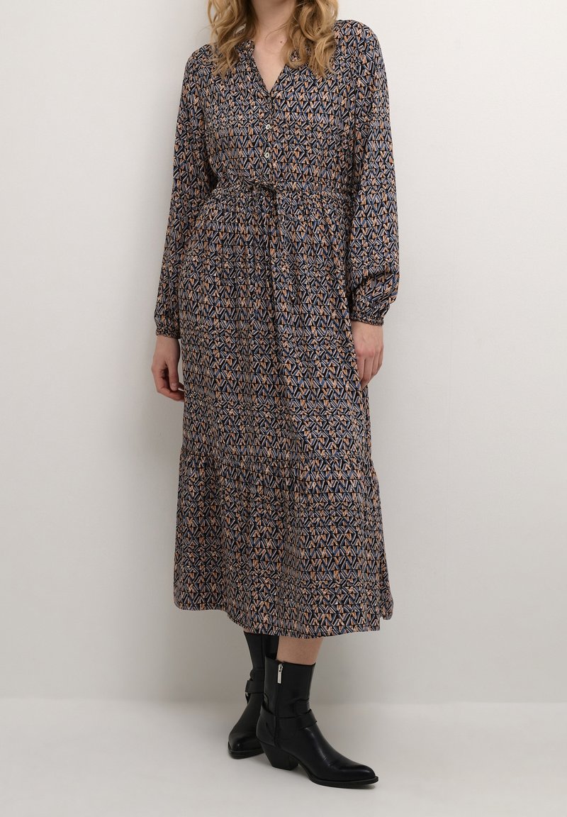 Femme portant une robe midi à manches longues avec un motif géométrique et une ceinture, associée à des bottes noires à talons mi-mollet, debout contre un mur uni.