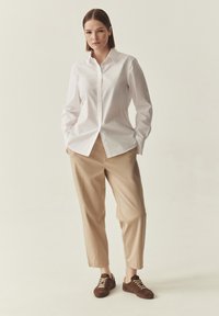 Chemise blanche à manches longues avec boutons, pantalon beige court et baskets en daim marron avec détails à lacets, sur fond neutre.