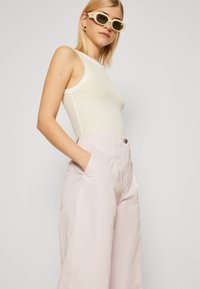 Débardeur blanc à côtes associé à un pantalon taille haute, large et rose clair. Modèle portant des lunettes de soleil claires, les mains dans les poches.