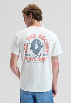 Mann iført hvit T-skjorte med animert vinylplate og tekst som promoterer en vinylbutikk for kjøp, salg og bytte av plater.