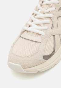 Sneaker beige con materiali in rete e suede, lacci bianchi, una parte superiore testurizzata e una suola in gomma chiara con accenti grigi.