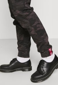 Mustat maastokuvioiset collegehousut, joissa on joustavat resorit lahkeissa ja sivutaskut, yhdistetty mustiin nahkakenkiin, joissa on valkoiset ompeleet, sekä valkoisiin sukkiin.