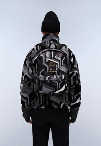Schwarzer und grauer Rucksack mit Muster, verstellbaren Riemen, Vordertasche und Logopatch. Das Material scheint glatt mit einer wasserabweisenden Beschichtung zu sein.