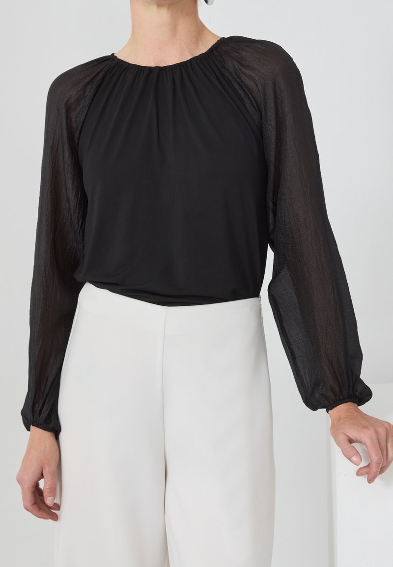 Femme portant un chemisier noir à manches longues transparentes et un pantalon blanc taille haute à jambes larges, debout avec une main posée sur une surface.