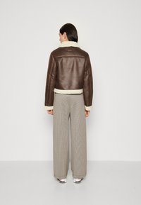 Giacca corta in pelle marrone con colletto e polsini in shearling crema, abbinata a pantaloni a quadri con ampia fantasia e sneakers bianche.